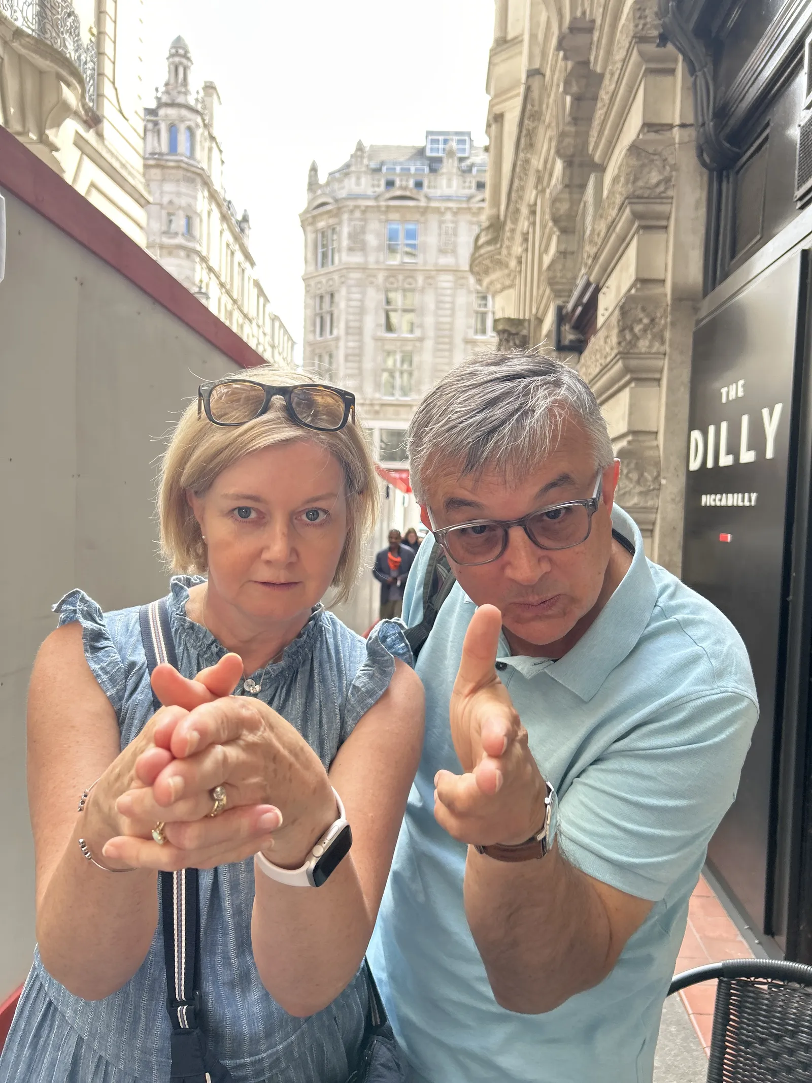 A woman and a man standing close together on a street with historical buildings in the background, both pointing their fingers forward as if holding imaginary guns in a playful pose; the woman wears a blue sleeveless dress, glasses on her head, a smartwatch, and the man wears a light blue polo shirt and glasses.