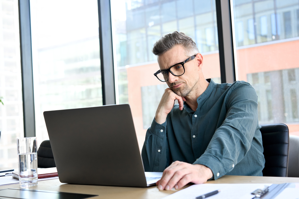 A man with glasses and a blue shirt sits in an office with large windows reviewing important information on his laptop.