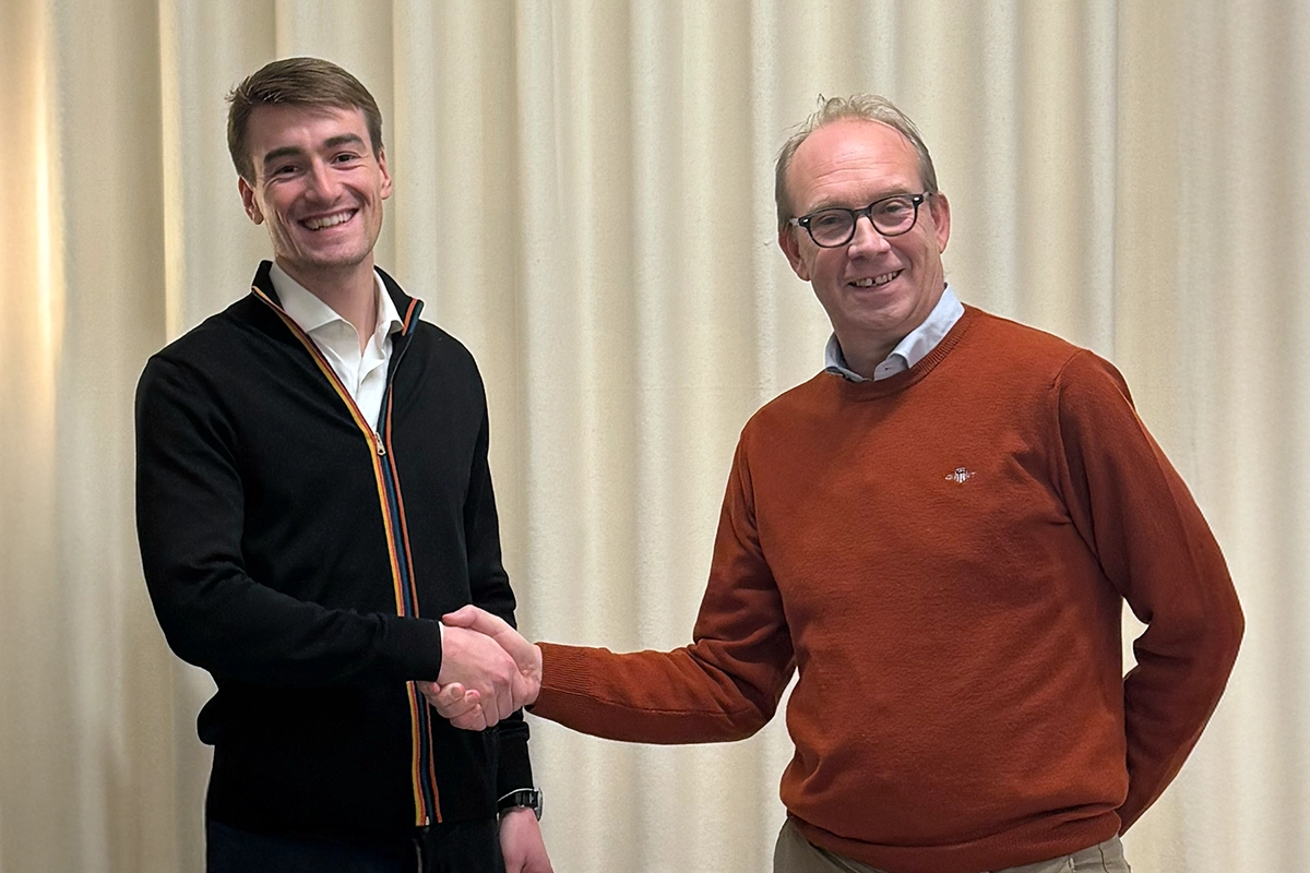 Two men smiling and shaking hands in front of a light-colored curtain.