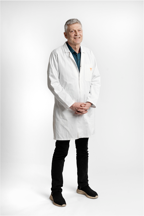 Middle-aged man with gray hair wearing a white lab coat over a dark shirt, standing with hands clasped in front of a white background.