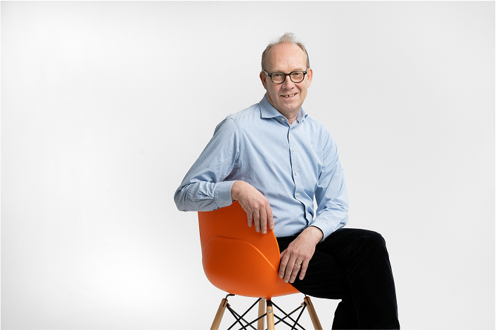 Middle-aged man wearing glasses and a light blue shirt sitting on an orange chair against a white background.