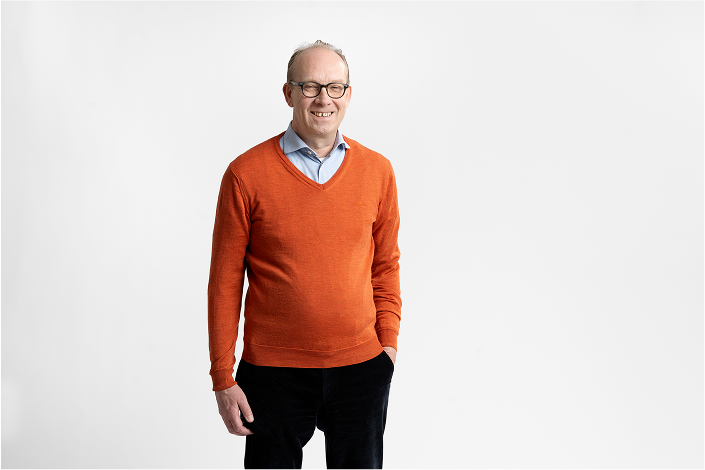Smiling man wearing glasses, an orange sweater over a light blue shirt, standing against a plain white background.