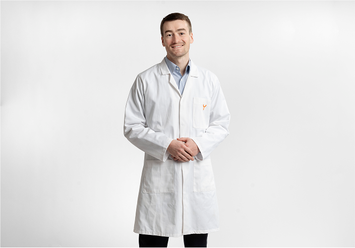Smiling male doctor wearing a white lab coat and light blue shirt standing with hands clasped in front against a plain white background.