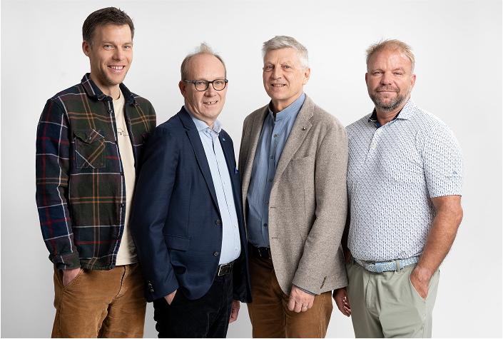 Four men standing side by side against a white background, smiling and dressed in casual and business casual attire.