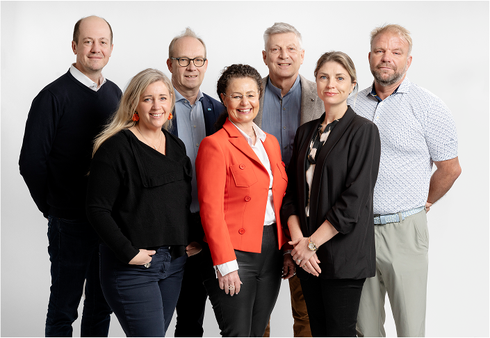Group portrait of seven professionally dressed adults standing and smiling against a white background.