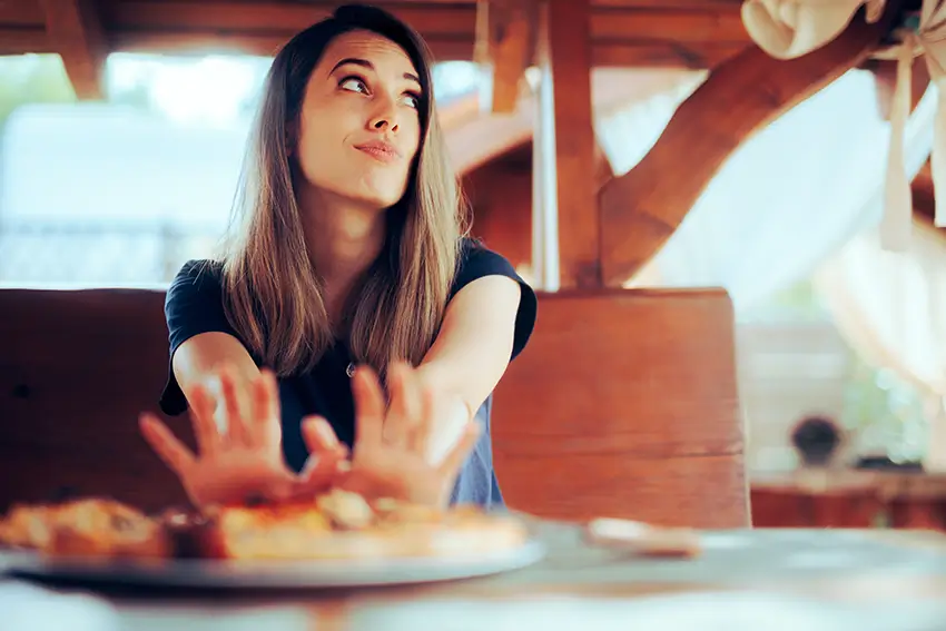 Young woman sitting at a table with a plate of food, pushing the plate away with her hands and looking upward thoughtfully.