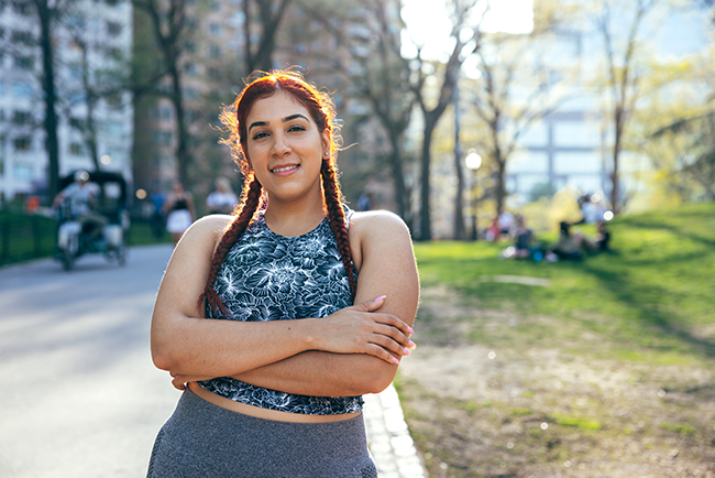 Smiling young woman with red braided hair and athletic wear standing with arms crossed in a sunlit park.