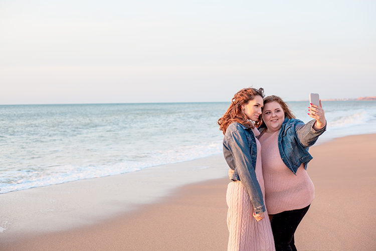 Two women on a beach near the ocean taking a selfie together during daylight.