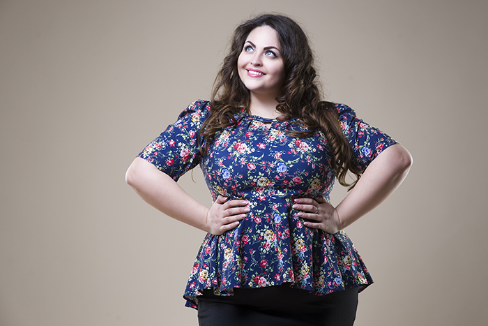 Smiling woman with long curly hair wearing a blue floral peplum top and black skirt, posing with hands on hips against a beige background.