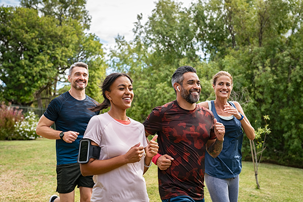 Four adults jogging outdoors on a grassy path surrounded by trees, smiling and wearing athletic clothes.