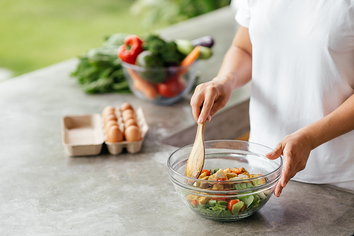 Person mixing fresh vegetable salad in a glass bowl on a kitchen counter with a carton of eggs and a bowl of assorted vegetables in the background.
