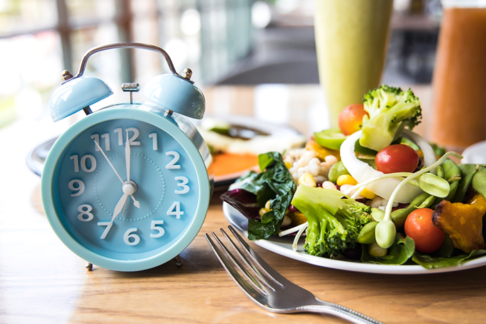 Light blue alarm clock showing 7:00 next to a plate of fresh salad and a fork on a wooden table.