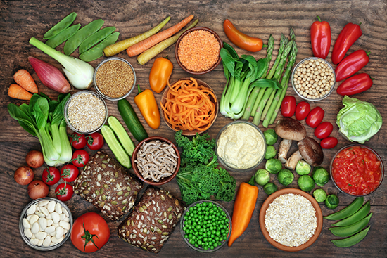 Assortment of fresh vegetables, legumes, whole grains, and seeds arranged on a wooden surface including carrots, asparagus, red bell peppers, green peas, tomatoes, leafy greens, lentils, oats, and whole grain bread.