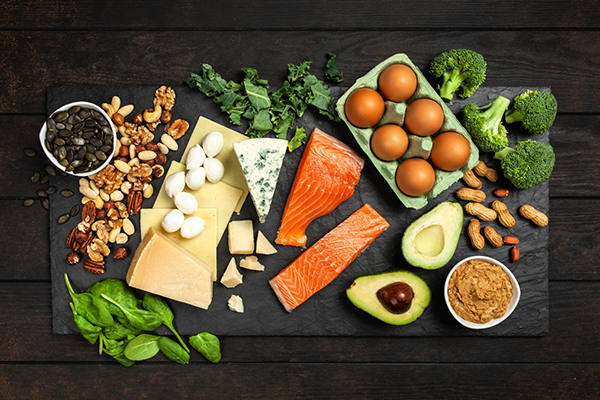 High-protein foods arranged on a black slate board including salmon fillets, avocado halves, eggs, various cheeses, nuts, spinach, broccoli, peanut butter, and seeds.