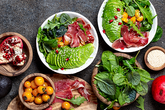 A selection of foods including meat, fish, eggs, avocado, leafy greens and other low-carbohydrate ingredients is displayed on a table. The image illustrates a ketogenic diet and highlights both ketosis and the potential risks and drawbacks associated with this way of eating.