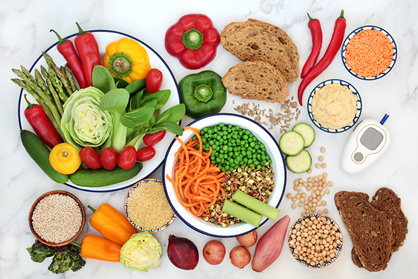 Variety of fresh vegetables, legumes, whole grain bread, and grains arranged on a white marble surface, with a glucose meter indicating a focus on healthy, diabetic-friendly foods.
