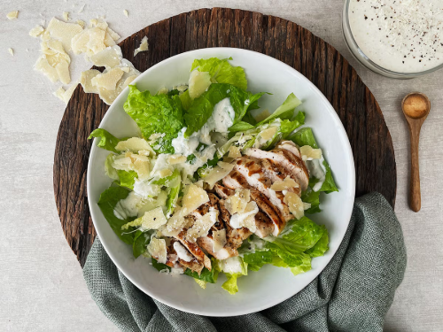 Grilled chicken Caesar salad with romaine lettuce and shaved Parmesan in a white bowl on a wooden board.