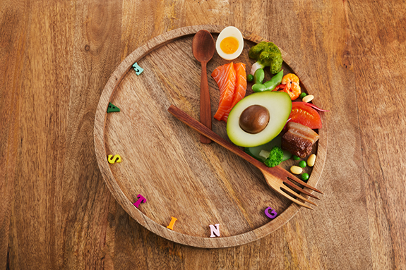 A healthy meal is arranged like a clock on a wooden table. The image illustrates fasting, meal timing and the potential drawbacks and challenges of intermittent fasting for weight management and overall health.