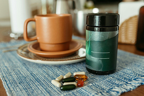 Assorted vitamin capsules and pills next to a dark green supplement bottle on a blue woven placemat, with a brown ceramic cup and saucer in the blurred background.
