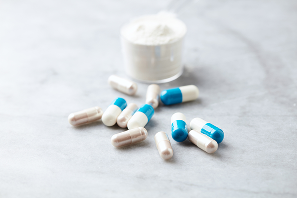 Blue and white capsules scattered near a transparent measuring scoop filled with white powder on a light surface.