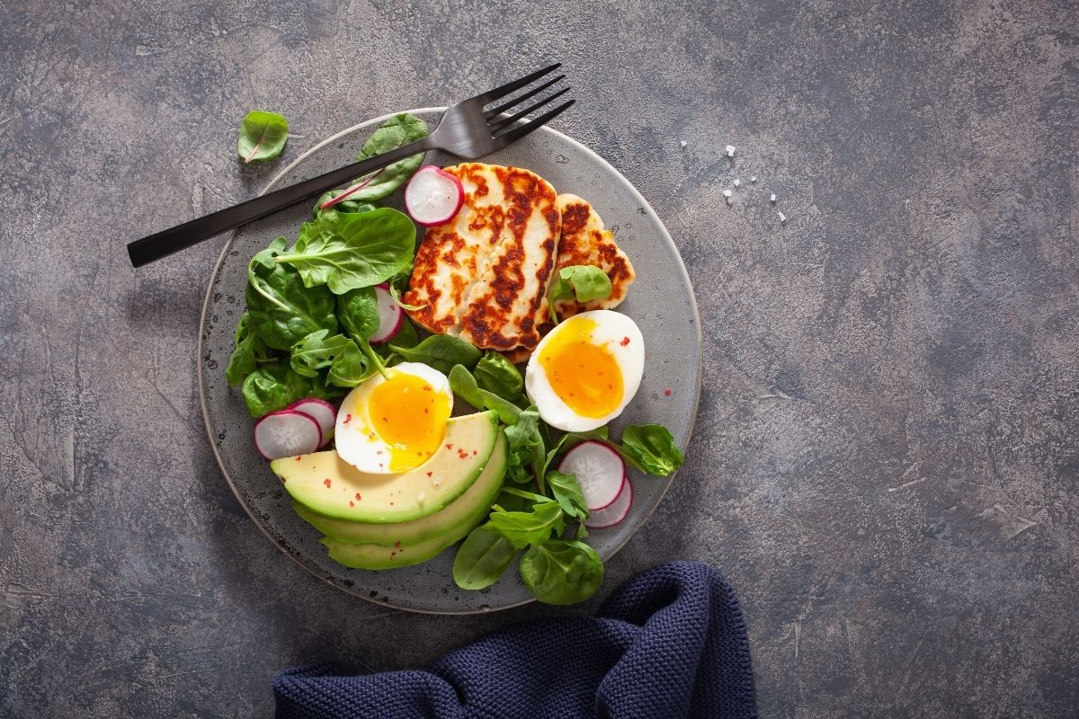 Plate with spinach leaves, sliced radishes, grilled halloumi cheese, two halves of a soft-boiled egg, and sliced avocado on a gray textured surface.