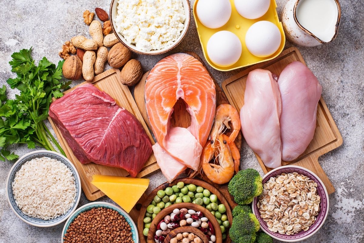 Variety of high-protein foods including raw beef, salmon, chicken breasts, eggs, shrimp, cottage cheese, nuts, cheese, rice, beans, peas, broccoli, oats, and milk on a textured surface.