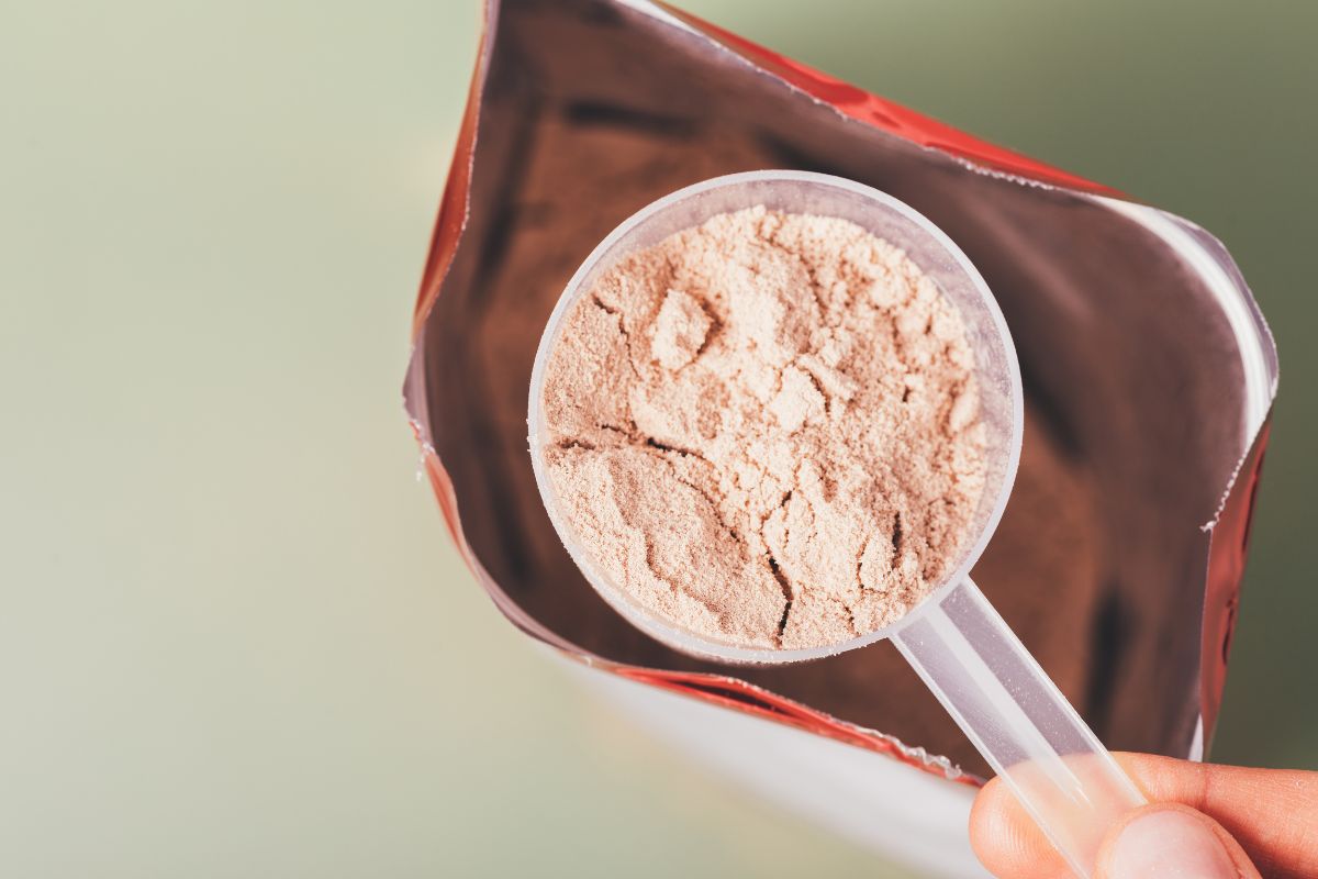 Hand holding a scoop of light brown protein powder over an open bag.