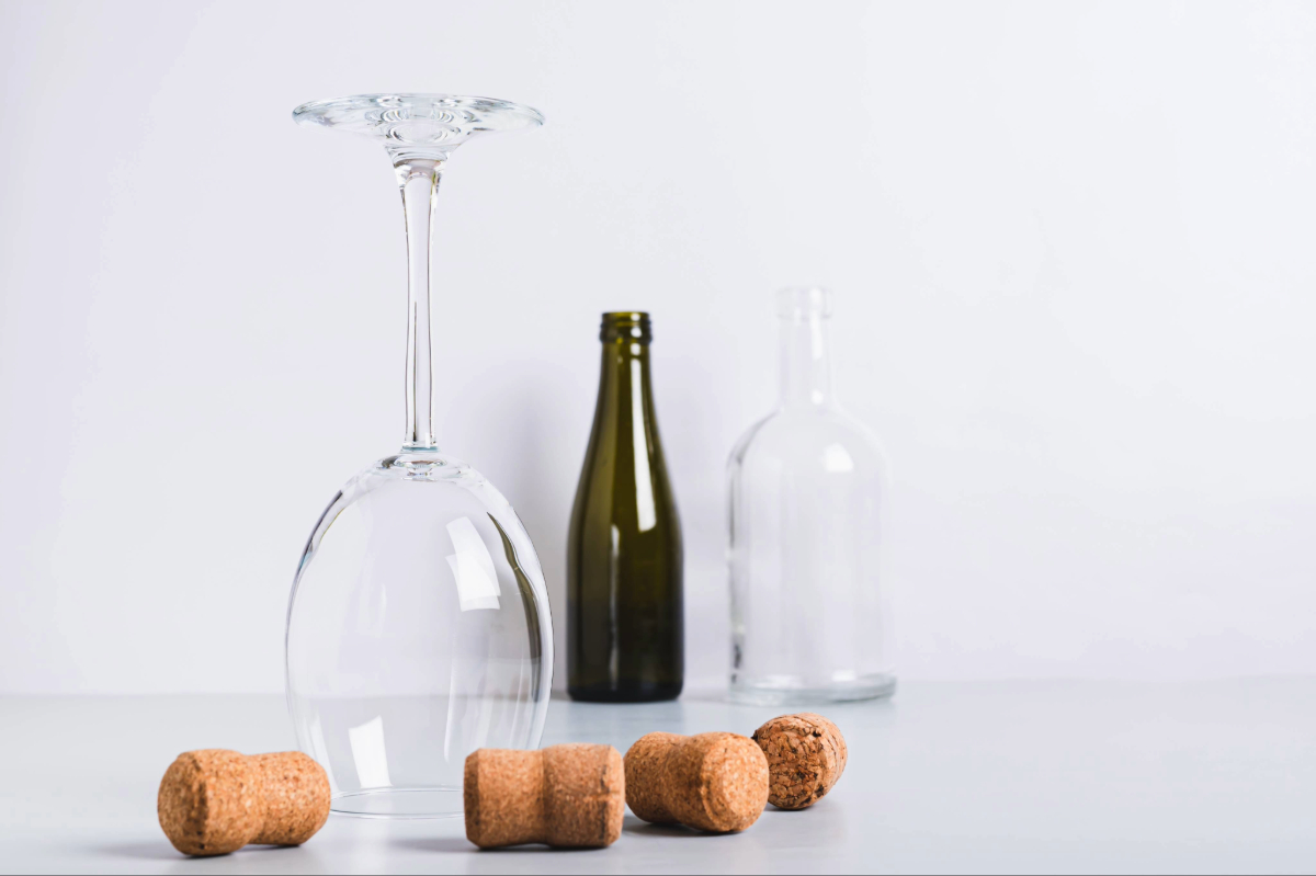 Upside-down wine glass with four corks and two empty wine bottles in the background on a white surface.