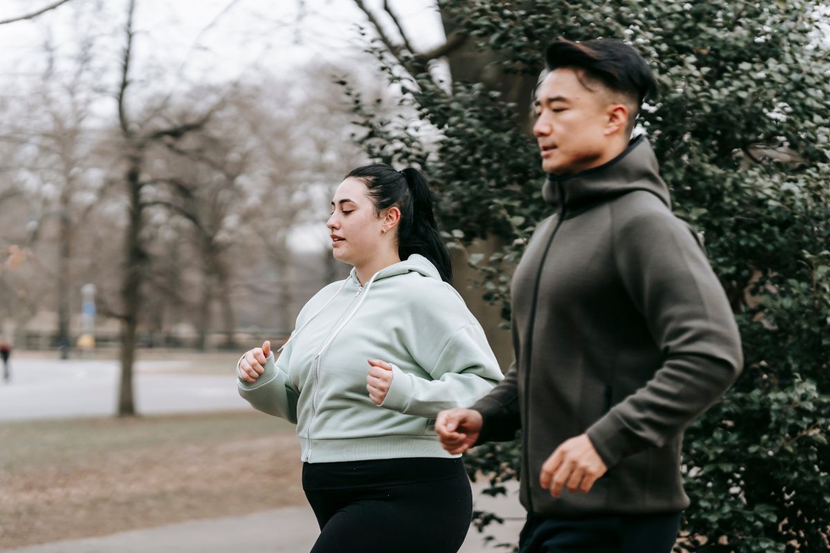 A man and a woman jogging outdoors on a pathway surrounded by trees and bushes in cool weather.