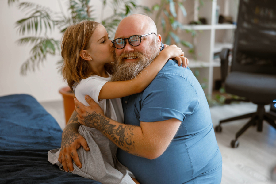 Bearded man with glasses hugging and smiling with a young girl who is kissing his cheek in a cozy indoor setting.
