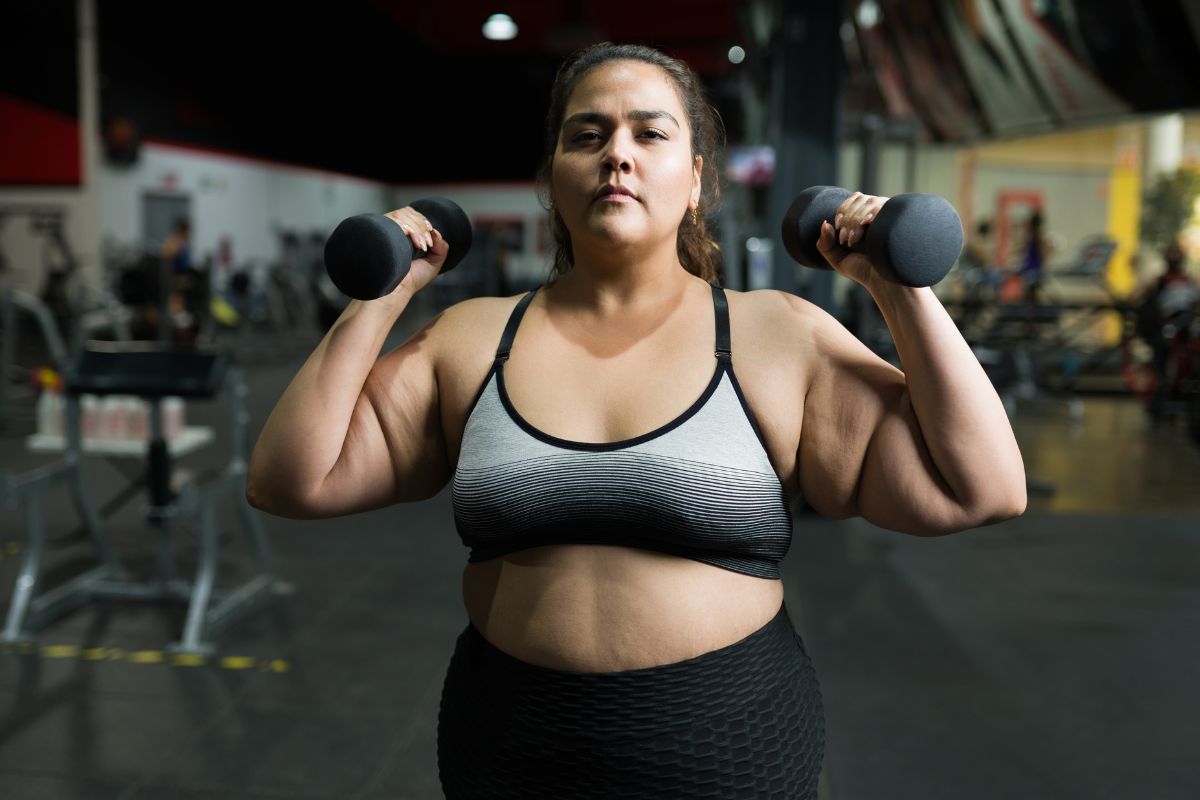 Confident woman in workout clothes lifting two dumbbells in a gym.