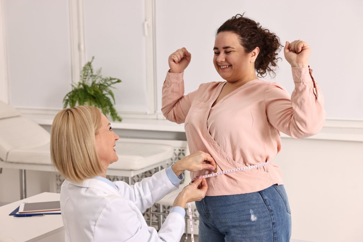 Smiling woman in pink blouse celebrating as a healthcare professional measures her waist with a tape measure in a medical office.