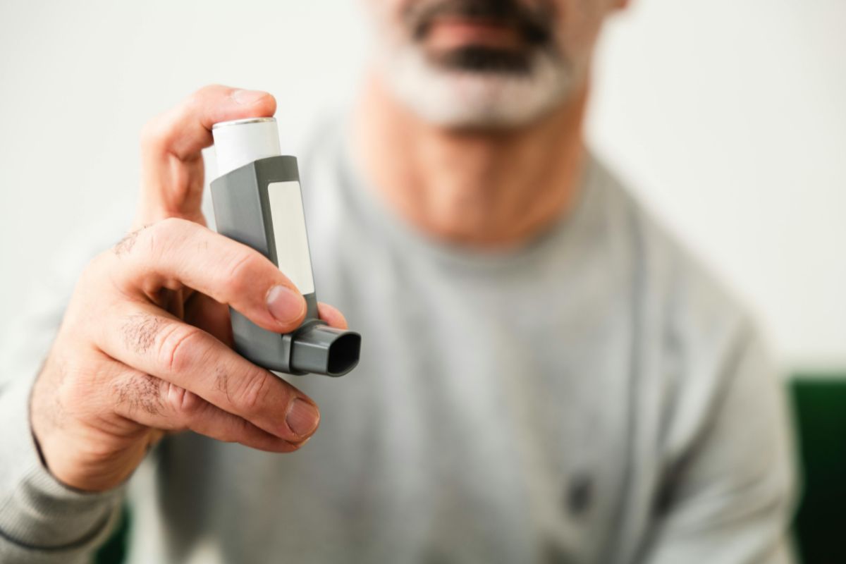 Close-up of a man holding a gray asthma inhaler toward the camera.