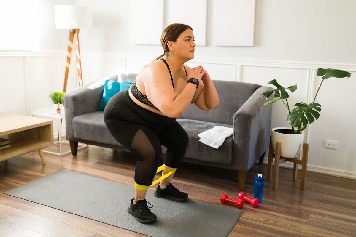 Woman exercising at home with a resistance band around her legs, doing a squat on a gray yoga mat.