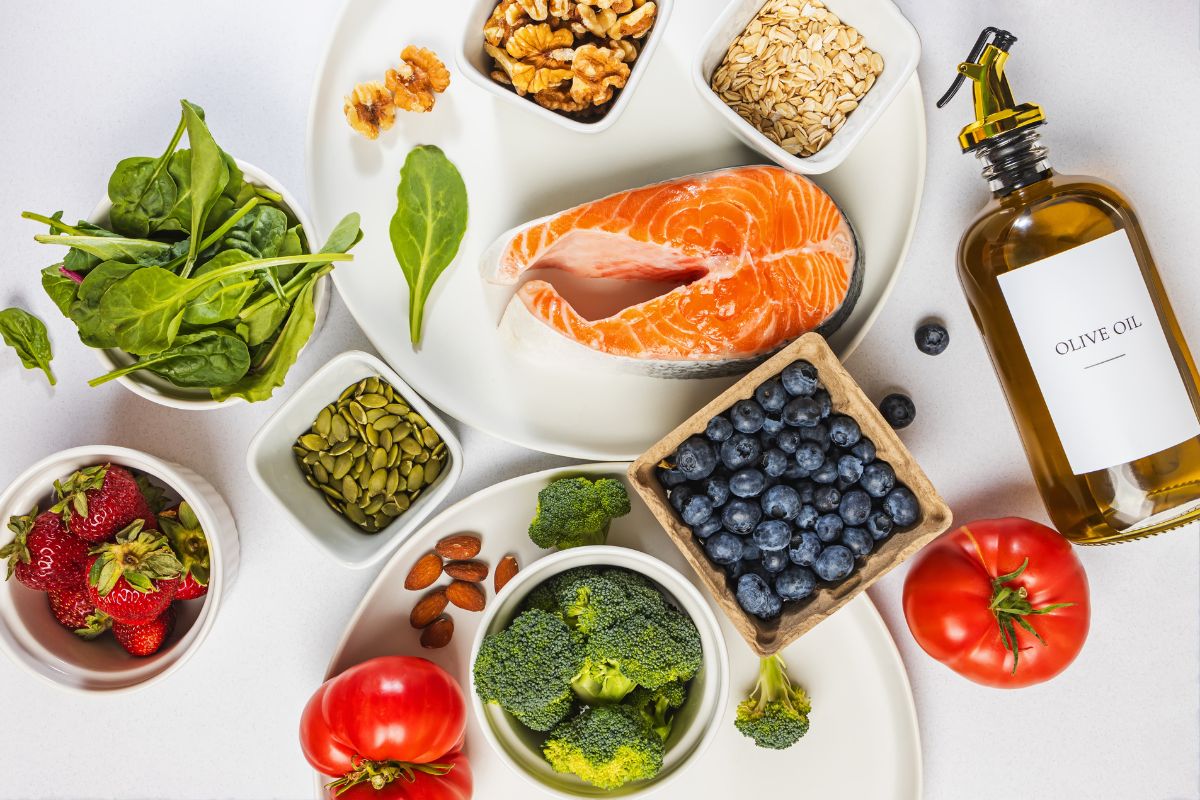 Assorted anti-inflammatory foods including salmon, blueberries, spinach, broccoli, strawberries, tomatoes, walnuts, almonds, pumpkin seeds, oats, and a bottle of olive oil.