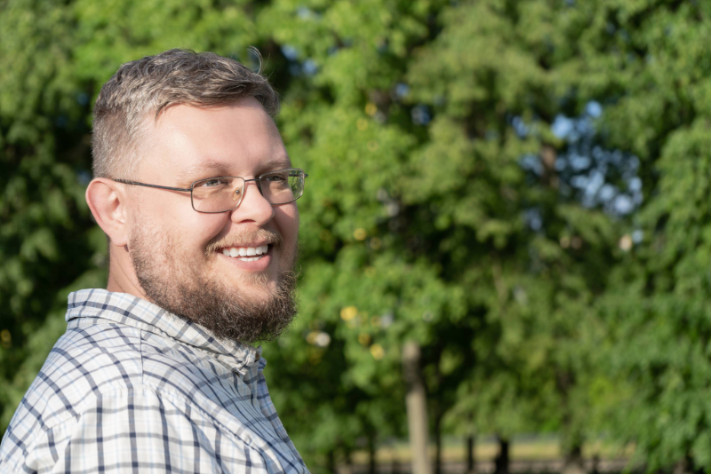Smiling bearded man wearing glasses and a plaid shirt standing outdoors with green trees in the background.