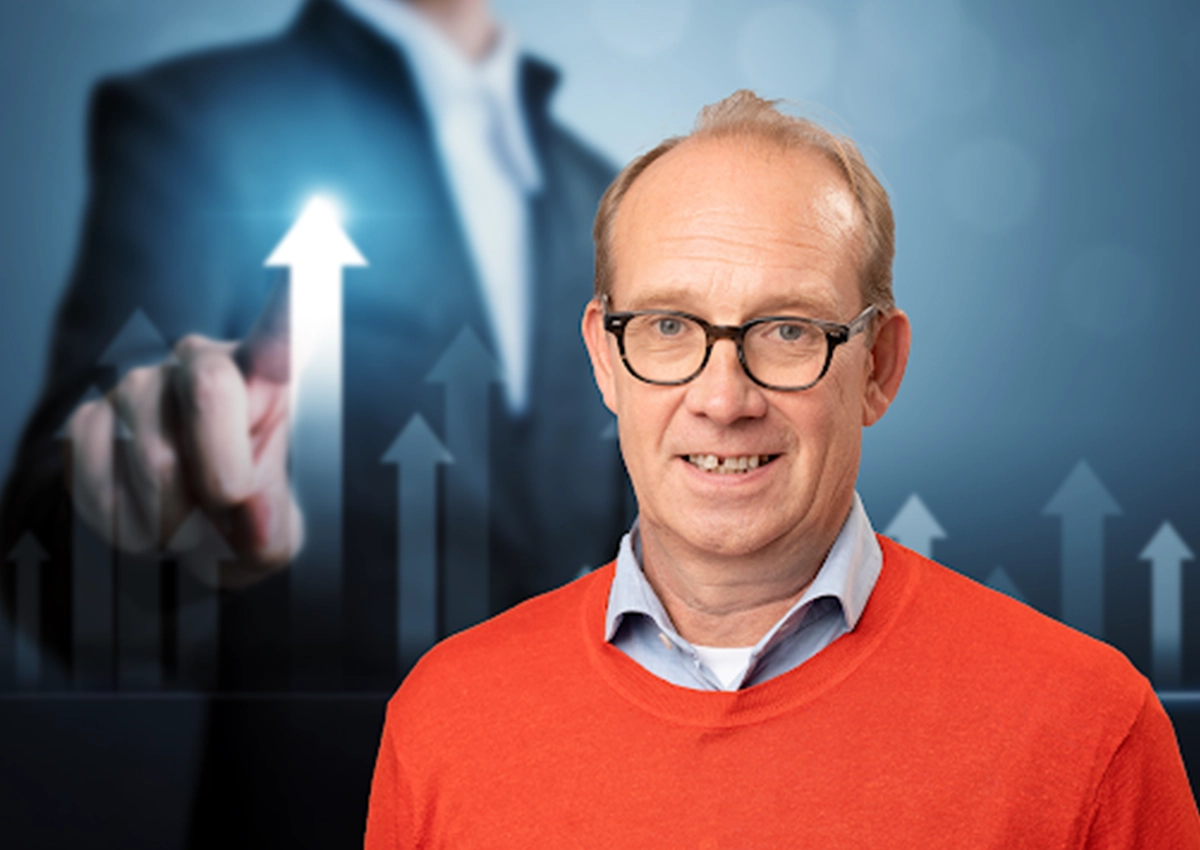 Smiling middle-aged man wearing glasses and an orange sweater with a blurred businessman pointing at upward arrows in the background.