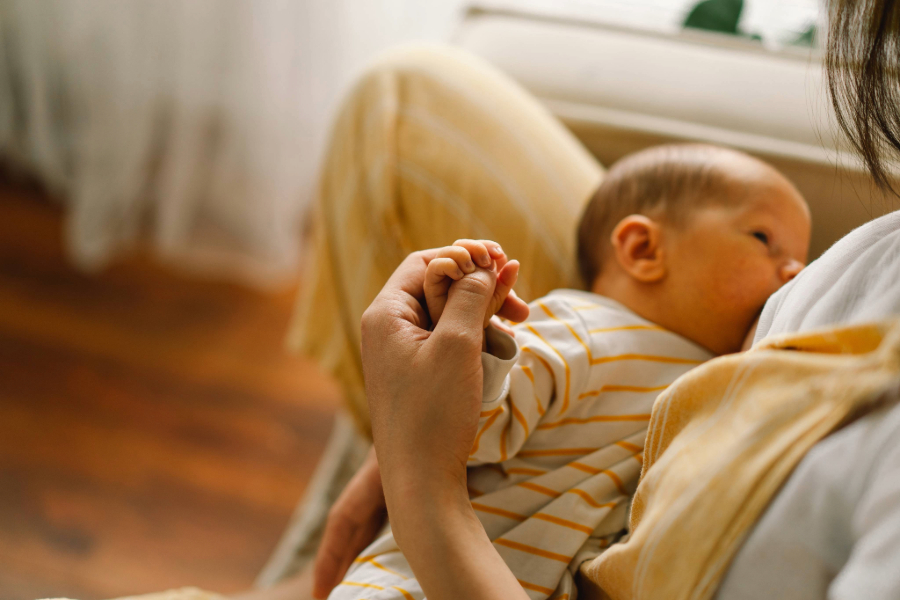 Close-up of a baby breastfeeding and holding an adult's finger while sitting on a lap indoors.