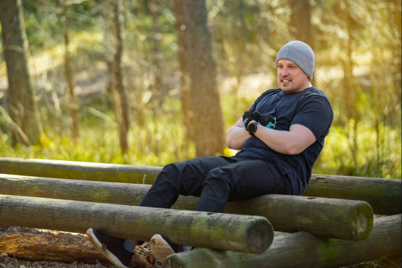 Man wearing a gray beanie and black sportswear sitting on wooden logs in a forest, smiling and exercising.