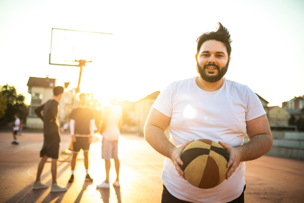 Man in a white t-shirt holding a basketball on an outdoor court during sunset with three other people standing near a basketball hoop in the background.