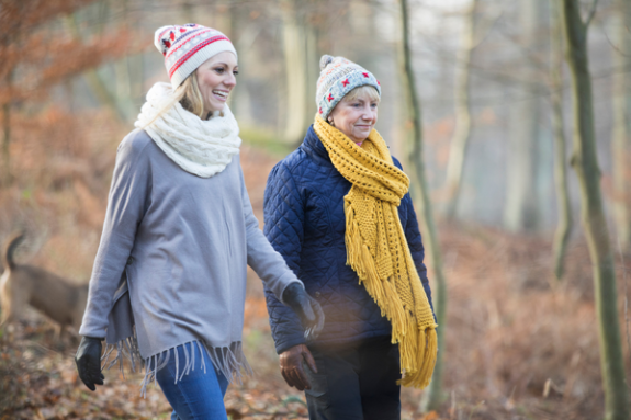 Two women walking in a forest during autumn wearing knitted hats, scarves, and gloves.