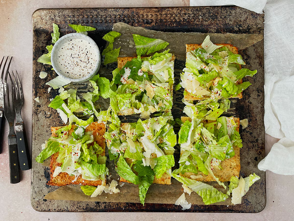 Flatbread cut into pieces topped with chopped romaine lettuce, shaved Parmesan cheese, and sprinkled chili flakes on a rustic baking sheet with a small bowl of creamy dressing and two forks nearby.