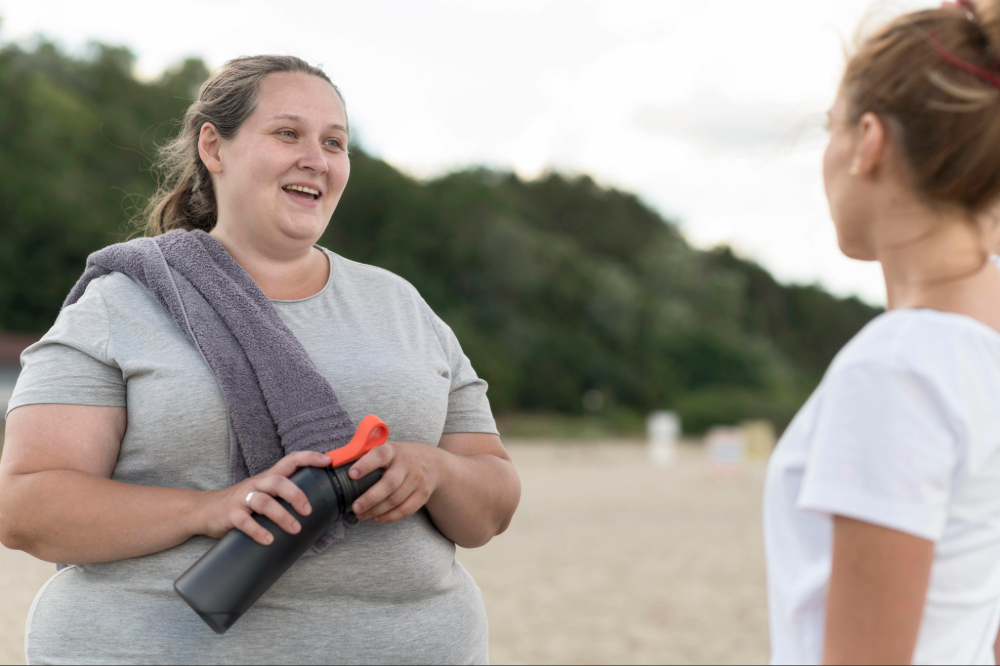 Two women outdoors, one holding a water bottle with a towel over her shoulder, engaged in conversation.