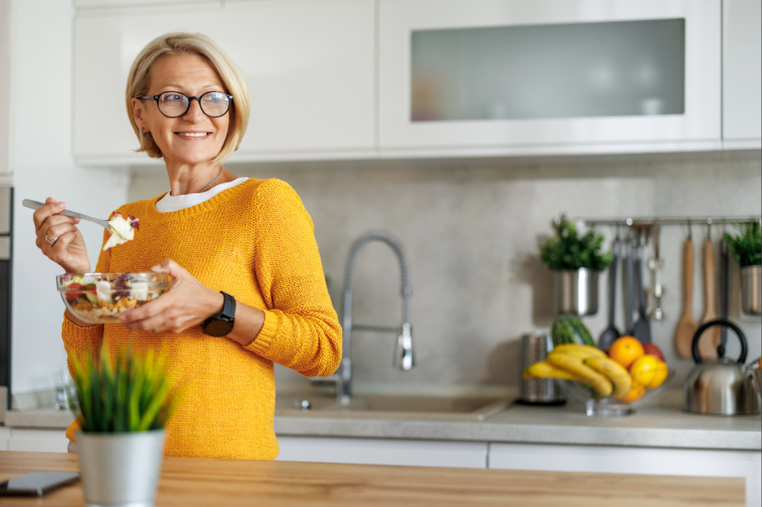 Smiling woman in yellow sweater eating a salad in a modern kitchen.