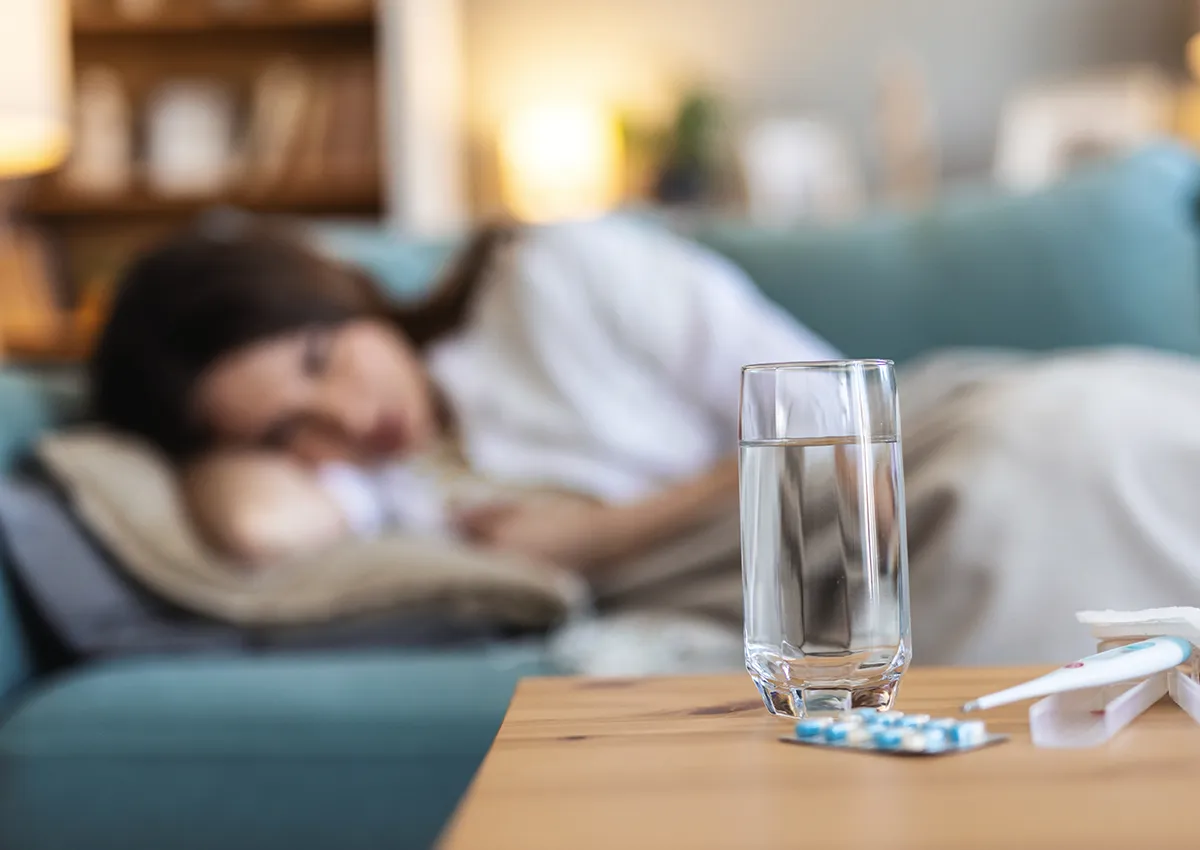 Glass of water, blister pack of pills, and digital thermometer on a wooden table with a woman lying on a couch in the background.