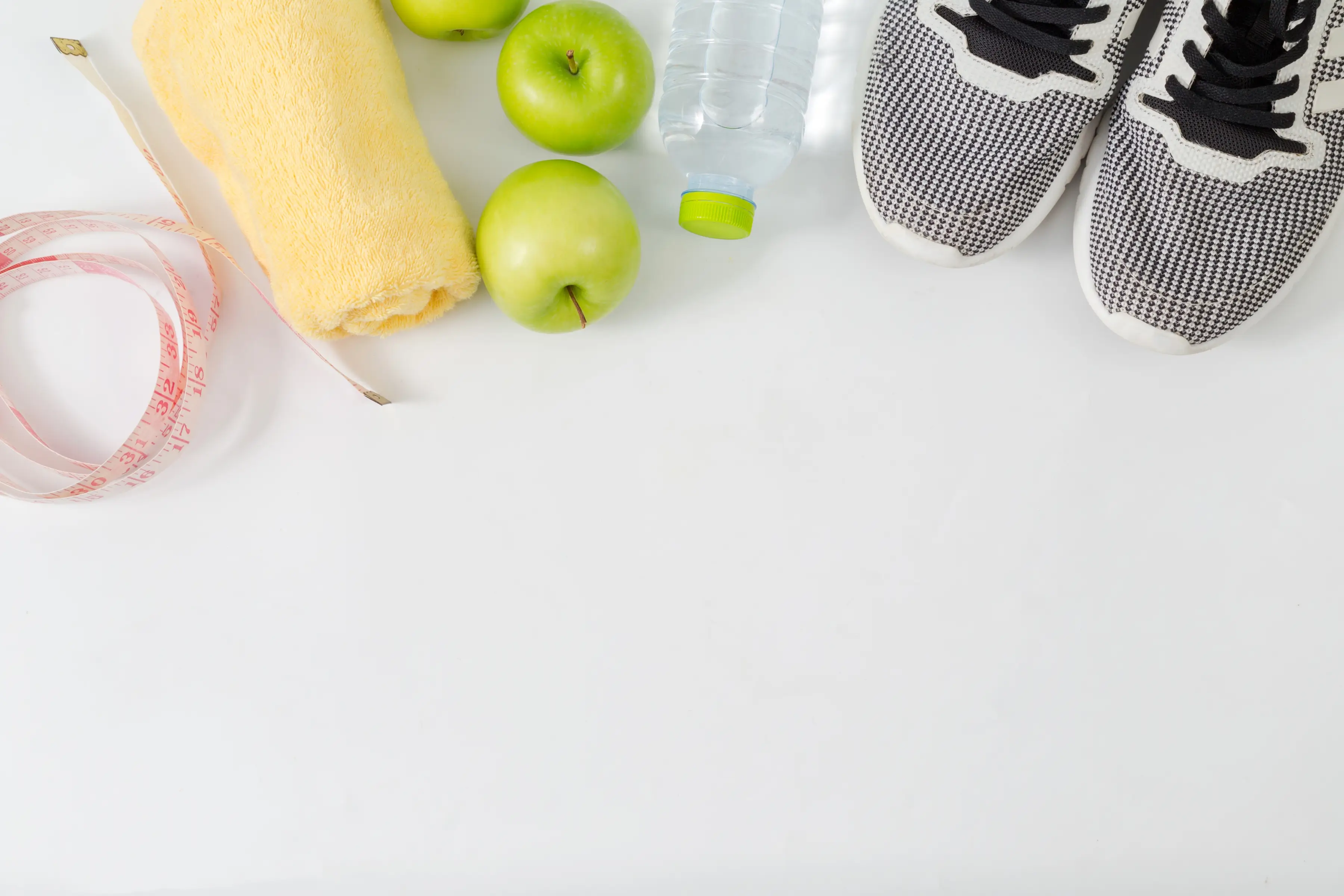 Flat lay of fitness items including a yellow towel, green apples, a water bottle, black-and-white sneakers, and a measuring tape on a white surface.
