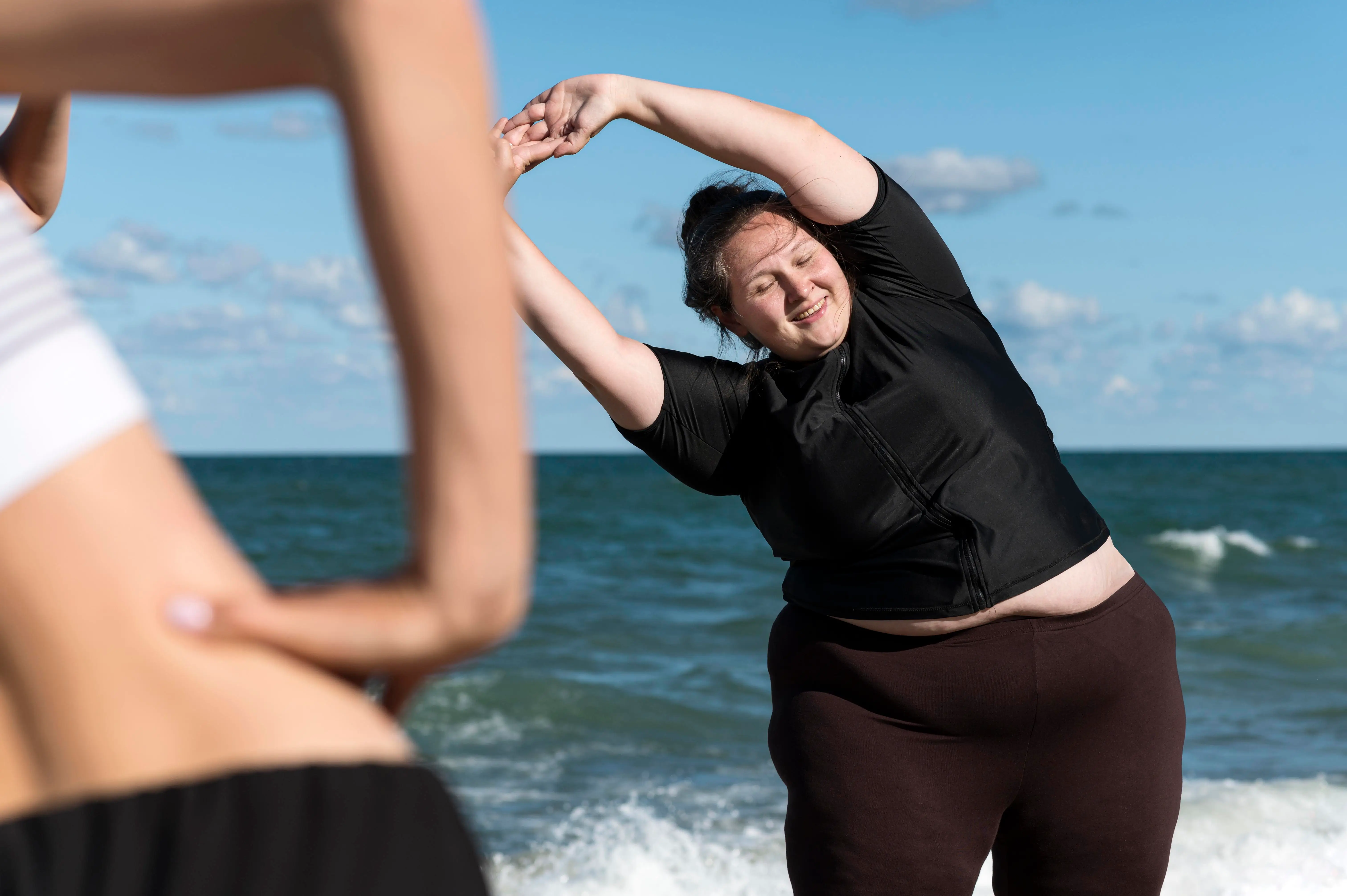 Two women stretching and exercising together on a beach by the ocean under a blue sky.
