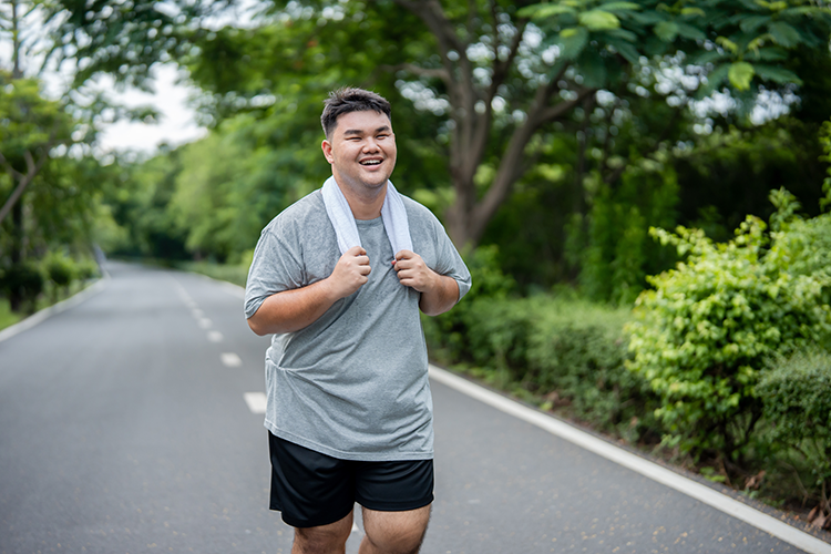 Smiling man in gray shirt and black shorts jogging outdoors on a tree-lined road with a towel around his neck.