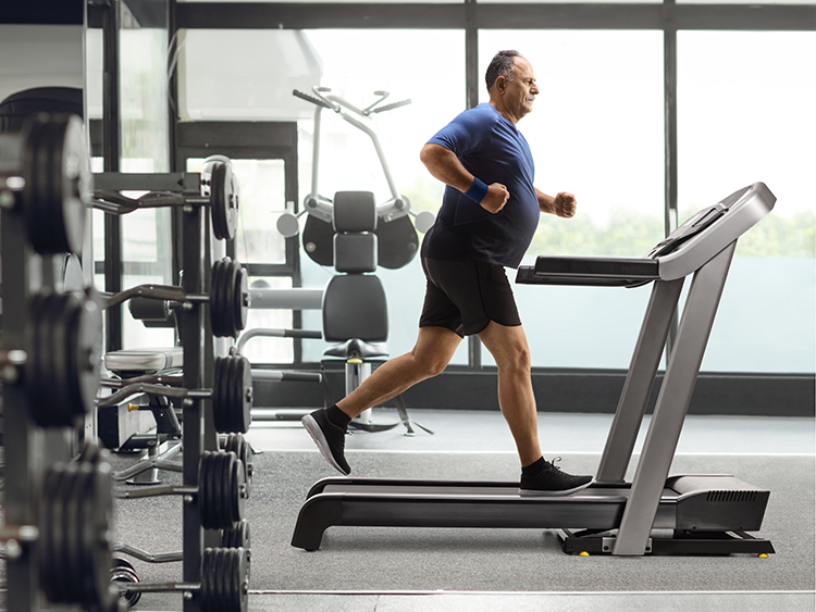 Middle-aged man running on a treadmill in a gym with weight equipment in the background.