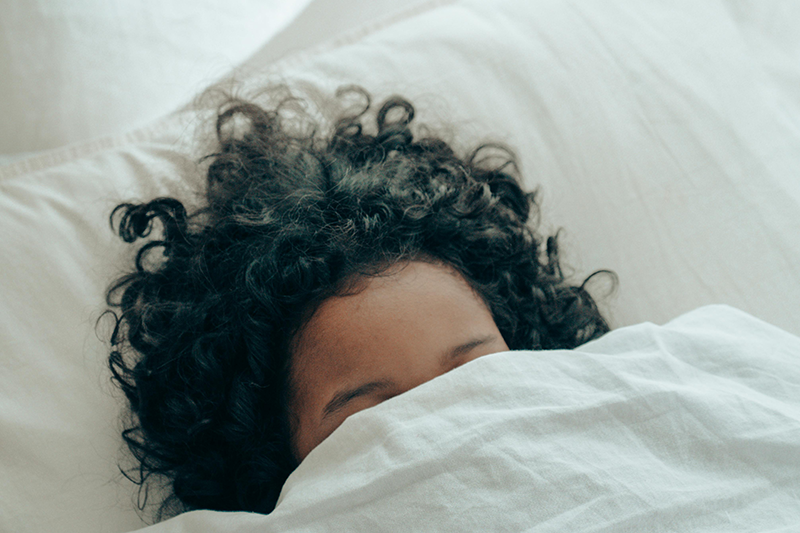 Child with curly hair partially hidden under a white blanket on a bed.